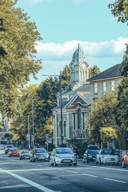 Street scene outside a residential property with a view of a historic town hall or church building featuring a clock tower with a spire, situated amid green trees and traditional brick houses. In the foreground, a driveway or loading area shows several parked cars and a few vehicles, possibly a small moving van or a vehicle used by Man with Van Southgate, involved in the home relocation process. The pavement is clear, and the weather appears bright with a blue sky and scattered clouds. Visible objects include wooden packing crates, cardboard boxes, and protective blankets, suggesting ongoing furniture transport and packing activities associated with house removals and moving services. The scene captures the typical logistics involved in loading and parking for a local removal or furniture transport, consistent with the service provided by Man with Van Southgate as part of a professional moving operation in Southgate.