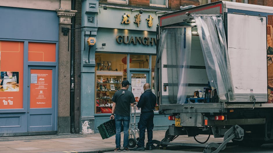 Two men are positioned on the sidewalk in front of a storefront named 'GUANGH' that features gold Chinese characters above the entrance. One man is wearing a black cap, black shirt, and jeans, and is 