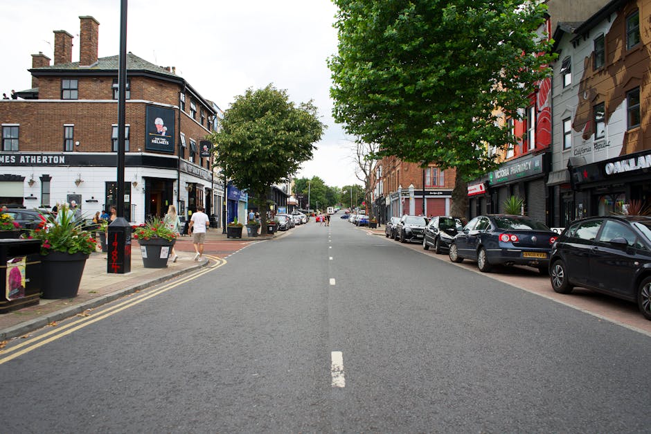 A view of a residential street on Bramley Road & Southgate Green, showing parked cars along both sides of the road, including sedans and hatchbacks, with some vehicles partially blocking the sidewalk. The pavement features decorative planters with flowers, and several pedestrians are walking along the sidewalk, some near the storefronts of local businesses such as cafes, pharmacies, and small shops. Green trees line the street, providing shade, and brick and painted buildings with windows and signage are visible, indicating a lively, urban environment. The scene appears to be during daylight with overcast weather, suitable for a house removal or furniture transport process. [COMPANY_NAME], specializing in removals, might be involved in a home relocation project in this area, where loading and unloading of furniture or boxes could take place nearby, although no moving activity is explicitly visible in this photo.