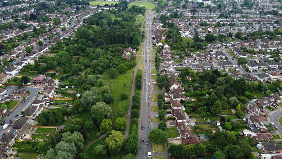 An aerial view of Bramley Road and Southgate Green in Southgate, showing a straight urban street lined with parked cars on both sides and residential houses with gardens. To the left of the road, there is a large, lush green park with dense trees, grass, and walking paths. Residential properties feature brick walls, driveways, and garden fences, with some houses having visible vehicles parked outside. The street appears active with a number of cars traveling in both directions, and the lighting indicates a daytime setting with natural sunlight illuminating the scene. The image captures the typical layout of a suburban area, with the greenery contrasting against the built environment, ideal for local house removals and furniture transport activities, as provided by Man with Van Southgate.