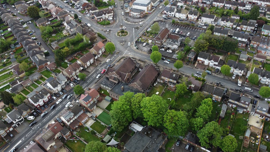 A view of a residential street on Bramley Road & Southgate Green, showing parked cars along both sides of the road, including sedans and hatchbacks, with some vehicles partially blocking the sidewalk. The pavement features decorative planters with flowers, and several pedestrians are walking along the sidewalk, some near the storefronts of local businesses such as cafes, pharmacies, and small shops. Green trees line the street, providing shade, and brick and painted buildings with windows and signage are visible, indicating a lively, urban environment. The scene appears to be during daylight with overcast weather, suitable for a house removal or furniture transport process. [COMPANY_NAME], specializing in removals, might be involved in a home relocation project in this area, where loading and unloading of furniture or boxes could take place nearby, although no moving activity is explicitly visible in this photo.