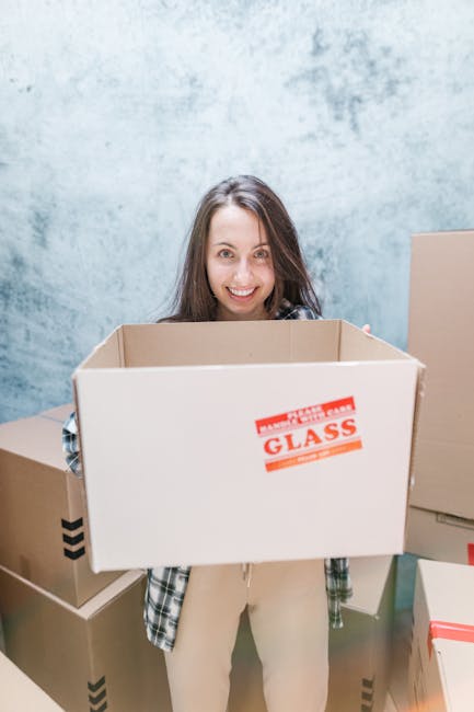 A young woman with long brown hair and a cheerful expression is standing indoors, holding a large empty cardboard box with a red and white 'glass' warning label on the front. She is dressed in a plaid shirt over a light-colored top, and stands surrounded by several other packed cardboard boxes of various sizes, some with black arrow symbols indicating the top side, in a space with a textured light blue wall in the background. The setting appears to be a home or storage area prepared for a house relocation or furniture transport, with the woman likely involved in packing or moving preparations as part of a home removal process. The scene emphasizes the packing and organizational stage of professional removals, such as those provided by Man with Van Southgate, during a home move or interim storage before transport.