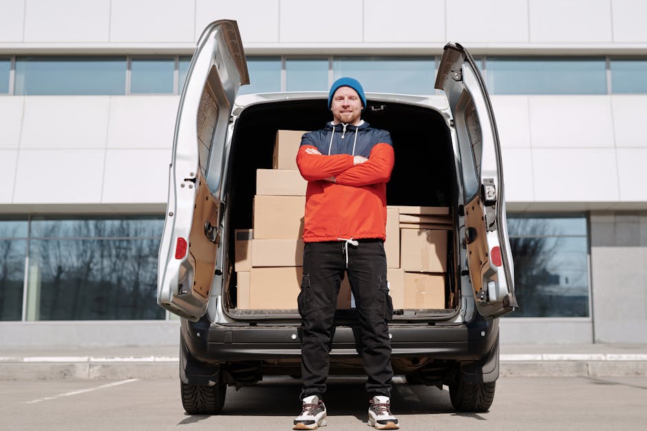 A man wearing a white t-shirt and jeans is standing on the pavement beside a white delivery van with its rear door open, engaged in packing or unpacking moving boxes made of cardboard, which are stack