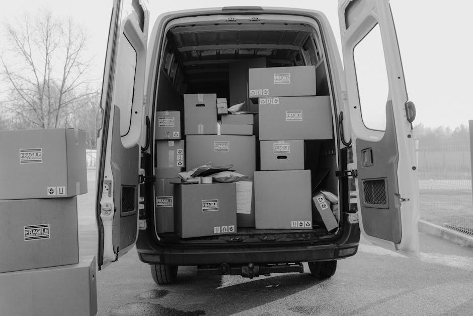 Photograph of the open rear doors of a white delivery van, filled with packed cardboard boxes of various sizes, some labeled with fragile handling symbols, and wrapped parcels arranged on the vehicle's floor and shelves. Additional boxes are stacked outside the van on the pavement nearby. The boxes contain household items ready for a home relocation, with some boxes secured with tape and plastic wrapping. The van is parked on a paved surface with a leafless tree and a fence visible in the background, indicating a residential or suburban area. The interior lighting highlights the organized loading process, which depicts furniture transport and packing details relevant to house removals. Man with Van Southgate provides this type of appliance and furniture loading service, as seen in the context of moving logistics and loading procedures for efficient household relocation.
