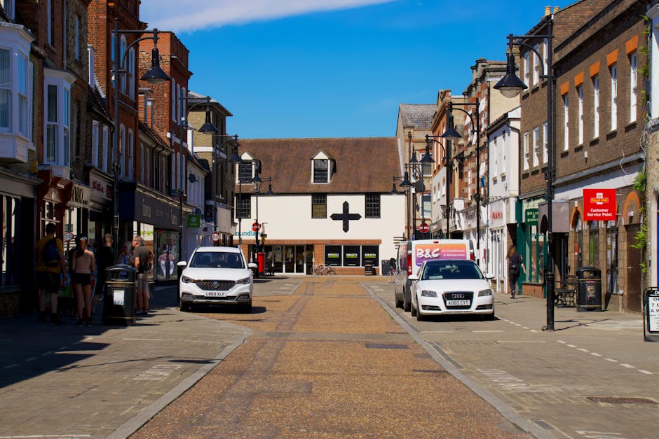 A view of a busy town centre street during daytime with parked cars, including a white van, and pedestrians walking on the sidewalks. On the left side, a group of people stand near a storefront, while on the right side, a white Audi car is parked next to a delivery truck. The street features a mixture of brick and stone paving, with lampposts lining both sides of the road. In the background, there is a white building with a black cross symbol, indicating a pharmacy or medical shop. The scene is well-lit with clear blue sky overhead, and the area appears to be suitable for home relocation or furniture transport when coordinating with [COMPANY_NAME] for efficient loading and moving services, especially within the context of Enfield Council Van Permits and loading rules as outlined on the Southgate page.