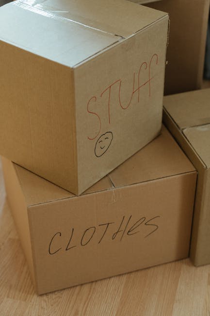 Close-up image of two cardboard moving boxes positioned on a wooden floor, with one box partially stacked on top of the other. The top box has the word 'STUFF' written in red marker on its side, accom