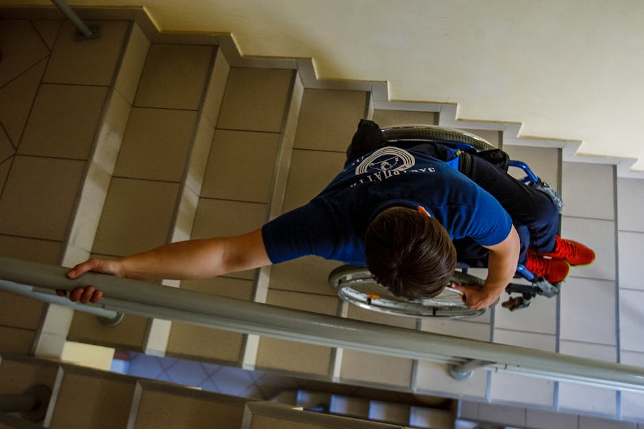 A man in a blue uniform, using a wheelchair with red socks and black hair, is navigating down a staircase inside a building. He is holding onto a handrail with his right hand while carefully descending the tiled stairs. The wheelchair is positioned on the staircase, indicating a horizontal loading or transport process, part of a home relocation or moving service. The staircase has beige tiles with white risers, and there is a yellow wall on the right side. Visible in the background are additional steps and a small portion of the floor at the top of the stairs. This scene illustrates the logistical challenge of moving furniture and belongings when stairs are narrow or lack a lift, a scenario where professional removals, such as those provided by Man with Van Southgate, are essential for safe transportation.