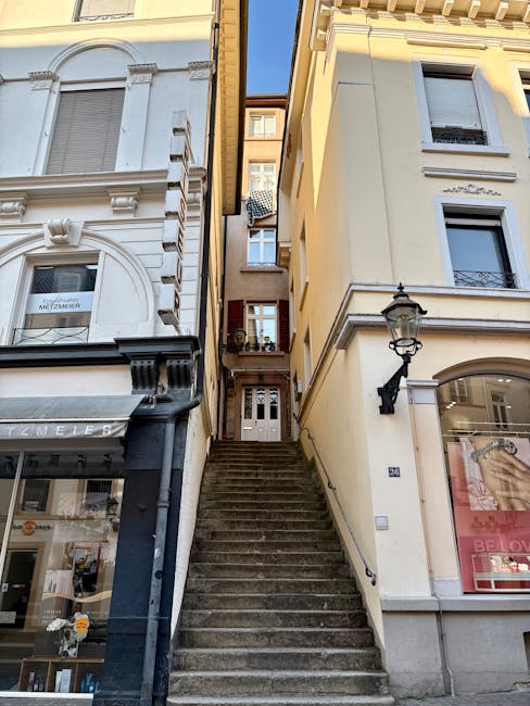 A set of outdoor concrete stairs leading up to an apartment entrance between two multi-story buildings in an urban setting. The left building features a white facade with decorative architectural details around the windows and a ground-floor shop with large glass windows displaying various items and a black awning. The right building has a cream-colored exterior with a black ornate street lamp mounted on the wall next to a window. At the top of the stairs, a small landing area shows a door opening into the apartment. The scene is lit by daylight, and the stairway appears to be part of a home relocation process involving furniture and boxes being carried or prepared for transport, with the presence of a man with a van service occasionally involved in loading or carrying belongings for an upcoming move, illustrating typical city-based furniture transport and packing procedures associated with professional removals like those offered by Man with Van Southgate.