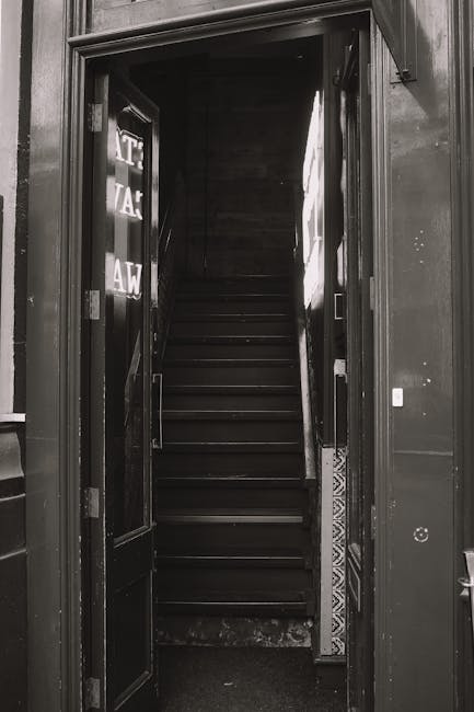 A man in a blue uniform, using a wheelchair with red socks and black hair, is navigating down a staircase inside a building. He is holding onto a handrail with his right hand while carefully descending the tiled stairs. The wheelchair is positioned on the staircase, indicating a horizontal loading or transport process, part of a home relocation or moving service. The staircase has beige tiles with white risers, and there is a yellow wall on the right side. Visible in the background are additional steps and a small portion of the floor at the top of the stairs. This scene illustrates the logistical challenge of moving furniture and belongings when stairs are narrow or lack a lift, a scenario where professional removals, such as those provided by Man with Van Southgate, are essential for safe transportation.