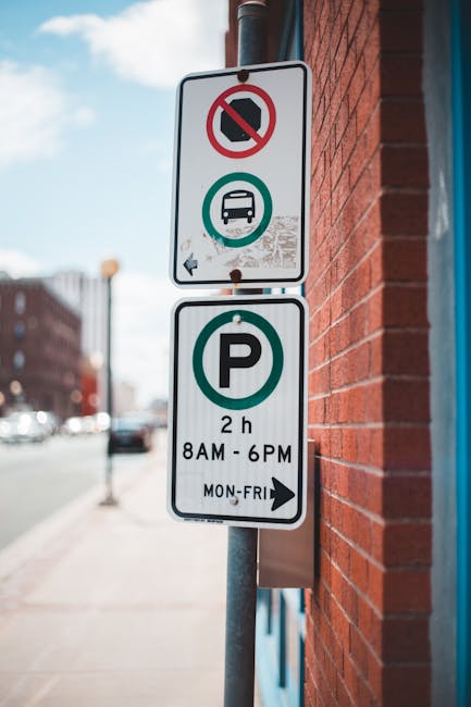 Photograph showing a vertical metal street sign mounted on a pole alongside a red brick building in Southgate, with a clear sky in the background. The top part of the sign has a white background with a red circle and diagonal line indicating no parking or stopping, along with a black van icon beneath, signifying restrictions for vehicle parking during certain times. Below, a green circle with a white 'P' indicates designated parking, specifying a maximum stay of two hours from 8 am to 6 pm, Monday to Friday. The sign is situated on a paved sidewalk, with moving traffic and parked cars visible in the background, highlighting urban street parking rules relevant for house removals or furniture transport in Southgate. The sign's clear visibility supports understanding parking regulations necessary for planning and executing property relocations in the area.