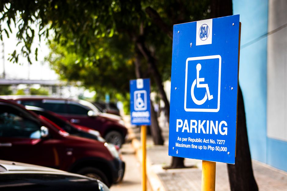 A blue parking sign with a white wheelchair symbol indicating accessible parking, mounted on a yellow post beside a paved parking area. The sign displays information about parking regulations under the Republic Act No. 7277, including a maximum fine of up to Php 50,000. In the background, there are several parked cars of various colors, including red and black, on the street. Behind the parking lot, there are green trees providing partial shade and a building structure with a blue wall. The scene is outdoors in daylight, and the image emphasizes parking and access considerations relevant to house removals or furniture transport services, as part of logistical planning for home relocation, handled by companies such as Man with Van Southgate.