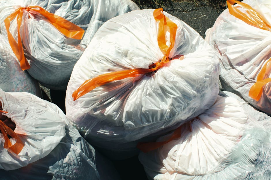 A close-up view of multiple large white plastic garbage bags filled with household waste, tied with orange plastic ties. The bags are stacked outdoors on the pavement near an open doorway, indicating they are ready for disposal or relocation. Some bags contain visible crumpled paper, cardboard, and other waste materials. The background shows part of a building's entrance with a dark ground surface, suggesting the waste is staged for collection or transportation as part of a house removal or moving process. The setting reflects typical packaging and waste disposal practices associated with home relocation or packing during a removal service by Man with Van Southgate, situated near a residential property.