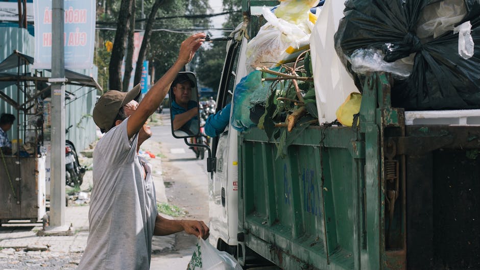 A man wearing a beige cap and light-colored shirt is seen standing beside a small green waste collection truck on a street. He is reaching up towards the truck's open cargo area, which is filled with various types of rubbish, including black and white plastic garbage bags, cardboard boxes, and green branches. The truck is positioned on the pavement near a sidewalk, with other parked motorcycles and a small structure visible in the background. The scene is outdoors during daytime, with trees and signage present. This image captures a waste disposal or collection activity, relevant to house removals or moving logistics, with Man with Van Southgate involved in the process of loading or unloading items.