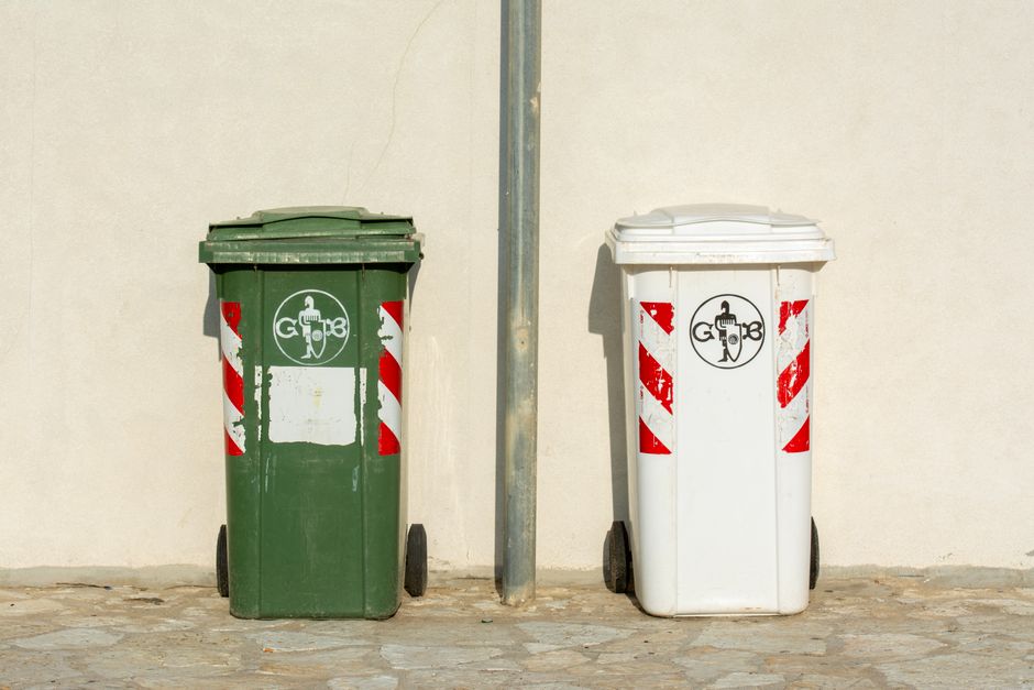 A close-up view of multiple large white plastic garbage bags filled with household waste, tied with orange plastic ties. The bags are stacked outdoors on the pavement near an open doorway, indicating they are ready for disposal or relocation. Some bags contain visible crumpled paper, cardboard, and other waste materials. The background shows part of a building's entrance with a dark ground surface, suggesting the waste is staged for collection or transportation as part of a house removal or moving process. The setting reflects typical packaging and waste disposal practices associated with home relocation or packing during a removal service by Man with Van Southgate, situated near a residential property.
