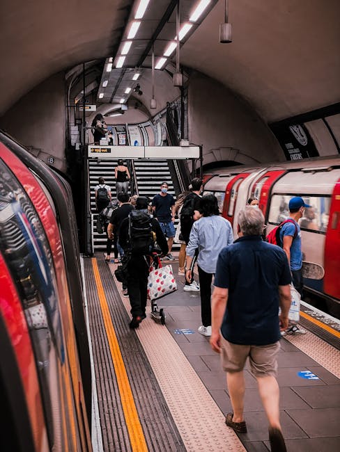 Inside Southgate Underground station, a red London Underground train is arriving or departing on the platform. The station features curved, tiled walls with white and black patterns, illuminated by overhead fluorescent lighting. A large blue advertisement for Man with Van Southgate is mounted on the wall beside the platform, related to house removals and transport services. Several white informational signs are visible on the wall, providing station and service details. The platform edge has a tactile yellow warning strip, and the floor is composed of light grey tiles. This setting reflects typical elements of a London underground station used for home relocation or furniture transport logistics, with trains serving as part of the moving process. The overall scene emphasizes the environment where household items and furniture might be loaded or unloaded during a house move, assisted by professional removals services.