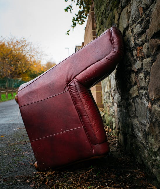 A red leather armchair has been placed outside against a stone wall on a pavement during daylight. The armchair is positioned at an angle, with the seat cushion and armrests visible, and appears to be in the process of being moved or discarded as part of a home relocation. Surrounding features include a brick wall and a background of trees with autumn foliage, along with an outdoor driveway or street area. The scene suggests a loading or unloading environment typical of furniture transport and packing logistics handled by Man with Van Southgate, demonstrating their interior furniture removal service as part of a house move.