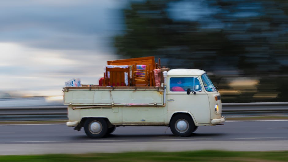 A vintage cream-colored van with a rounded front and small windows, loaded with household furniture and packing materials, is driving along a road during daytime. The cargo includes wooden chairs, a small table, and a cardboard box, all secured on the open bed of the vehicle. The items are covered with protective blankets and plastic wrapping to prevent damage during transit. The moving load indicates an active home relocation process involving furniture transport, with the van belonging to Man with Van Southgate engaged in a house removals service. The background features a blurred landscape of trees and sky, emphasizing the vehicle's motion and transportation of household belongings during a residential move, reflecting the logistics of packing and moving services typical of a professional removal company.